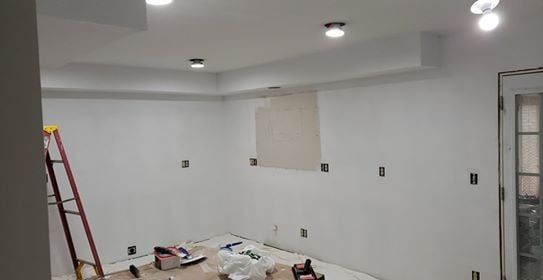 Interior under construction with white walls, recessed lighting, and electrical outlets. A ladder is present.