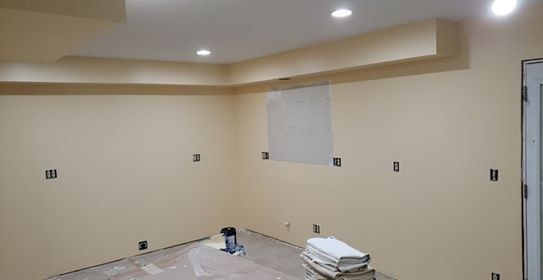 A room with beige walls and ceiling, electrical outlets, a small window, and some construction materials.