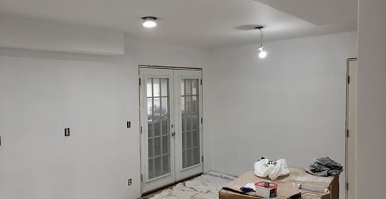 White-painted interior, French doors, overhead lights, and a table with supplies, suggesting a room under renovation.