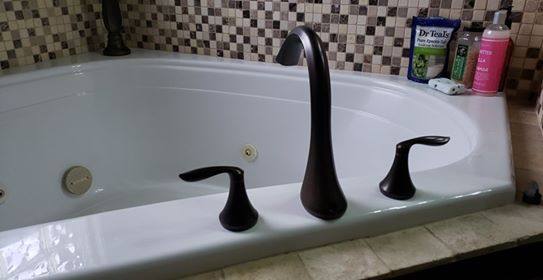 A black faucet and handles on a white bathtub, against a mosaic-tiled wall.