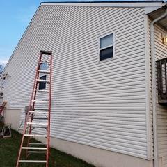 A tall orange ladder leans against a light-colored house with white siding, a small window, and a blue sky.