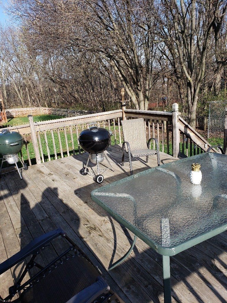 Deck with black grill, glass-top table, and wooden railing, surrounded by trees on a sunny day.