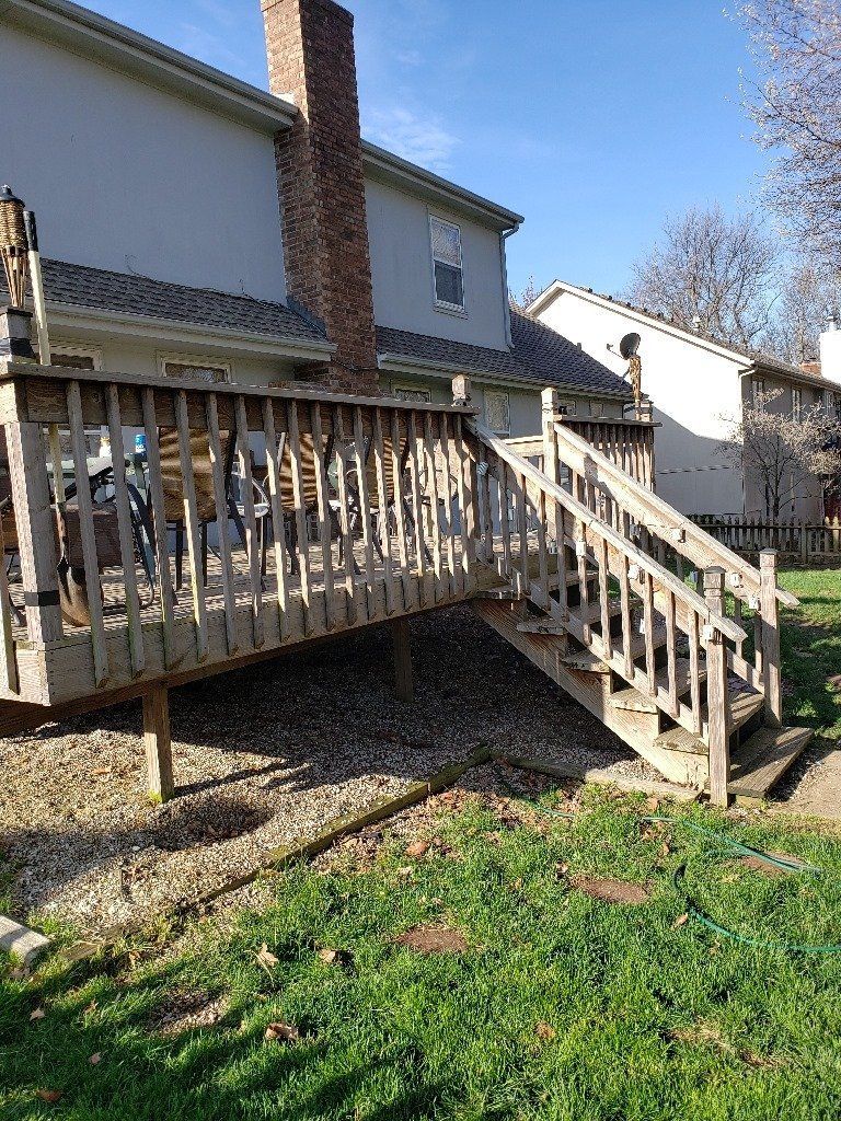 Wooden deck with stairs attached to a two-story house, surrounded by grass and gravel on a sunny day.