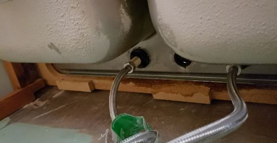 Underside view of a double sink with flexible supply lines connected. Water is visible, and wood framing is present.