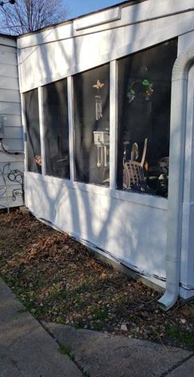 White screened-in porch on a house with plants and items visible inside; the ground has dried leaves.
