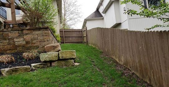 Backyard with a wooden fence and stone retaining wall. A sloped lawn leads to a wooden gate.