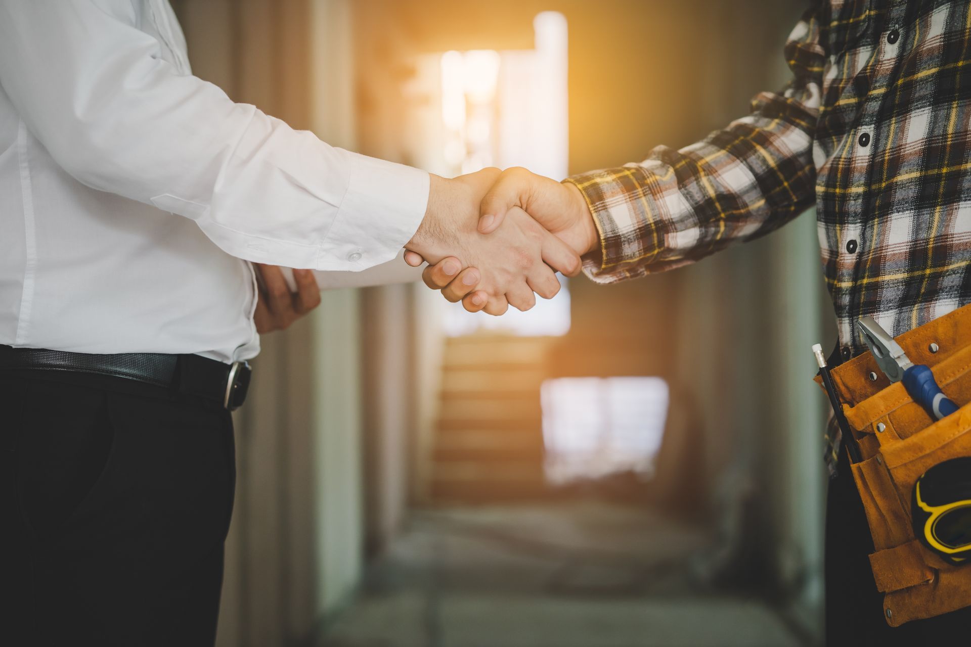 Two men shaking hands in a construction site; one in a white shirt, the other wearing plaid and a tool belt.