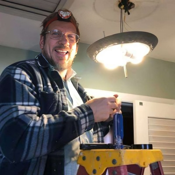 Man smiling, standing on a stepladder while working on a light fixture indoors. He wears a headlamp and flannel shirt.
