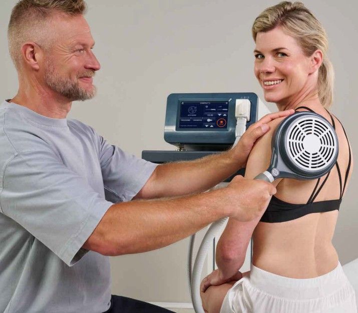 Therapist treating patient's shoulder with medical device. Both smiling in clinic.