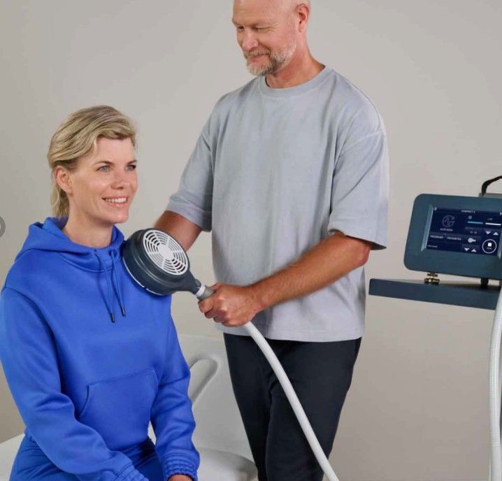 Man holding device to woman's neck, likely for therapy. Both are indoors, with a control panel visible.