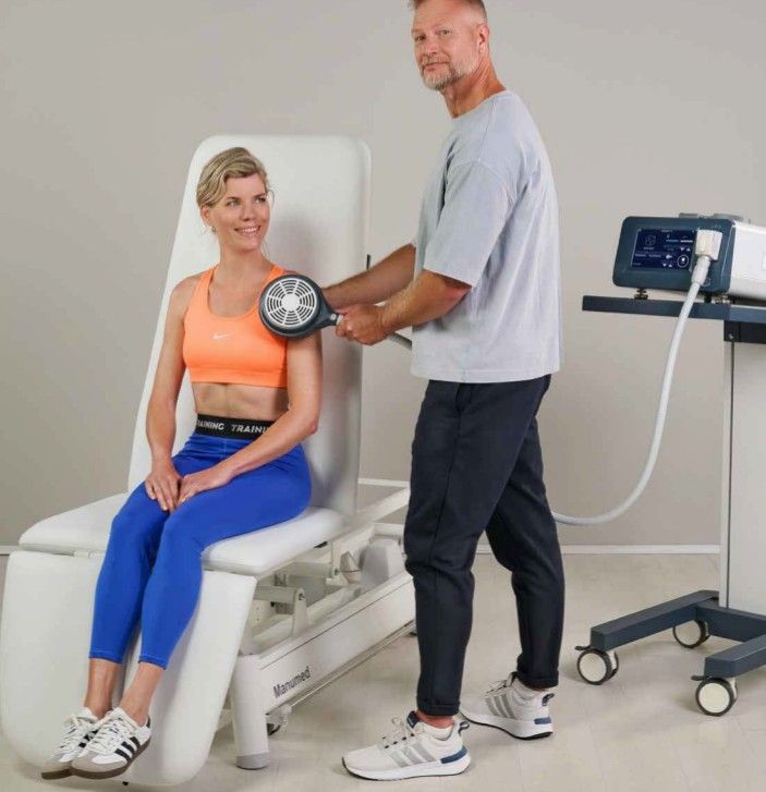 Therapist using a device on a seated person's shoulder. Light-skinned individuals in a clinical setting.