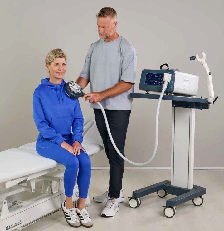 Man using a medical device on a woman's shoulder. Device on a wheeled cart in a clinic.