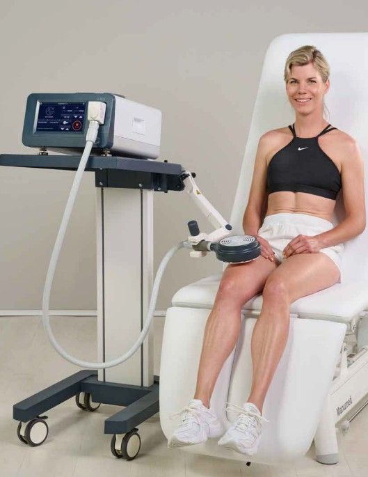 Woman receiving therapy treatment on her knee with a medical device. She sits on a medical table, smiling.