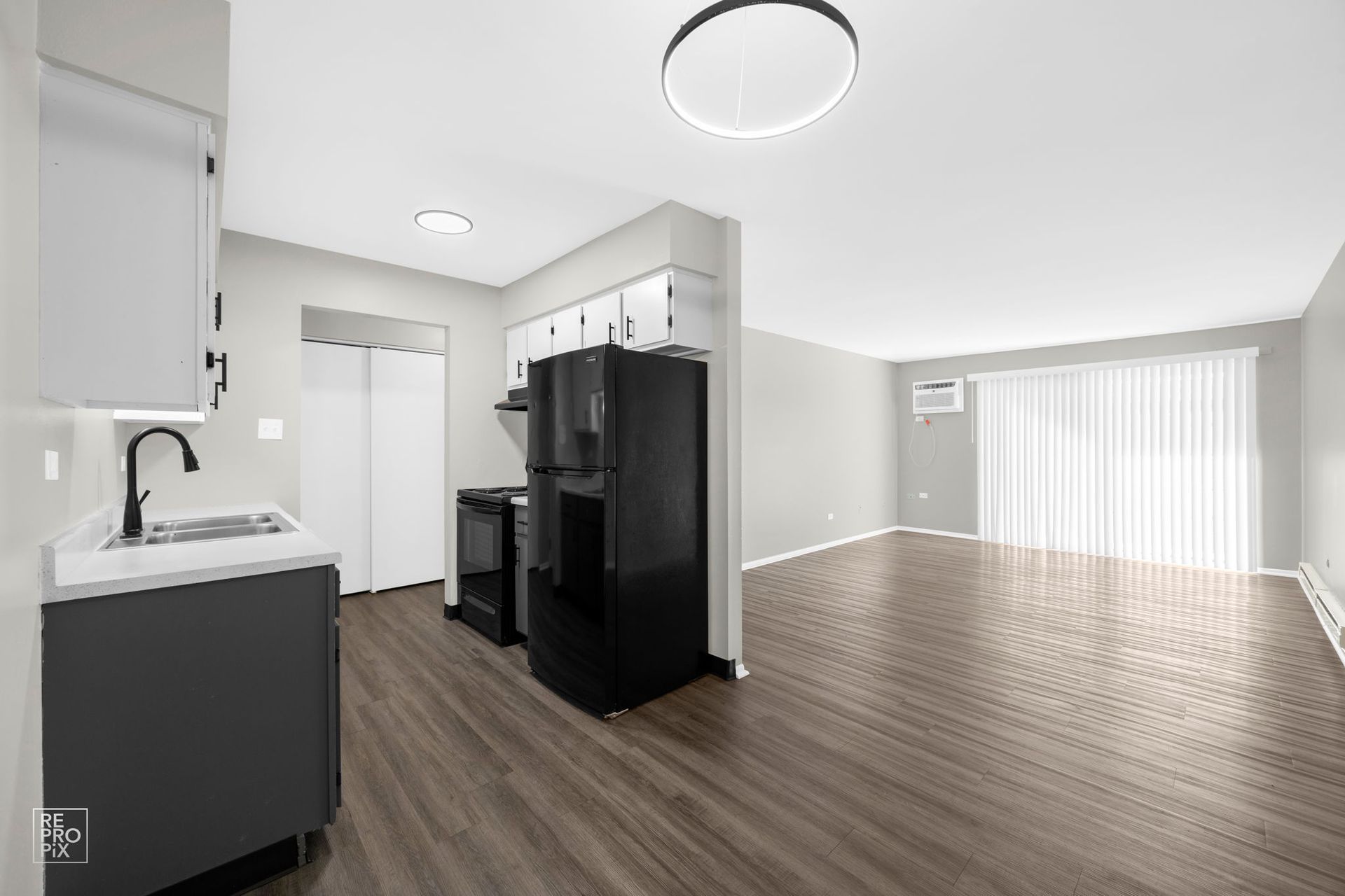Kitchen with gray cabinets, black appliances, and laminate flooring, opens to a bright living room with large windows.