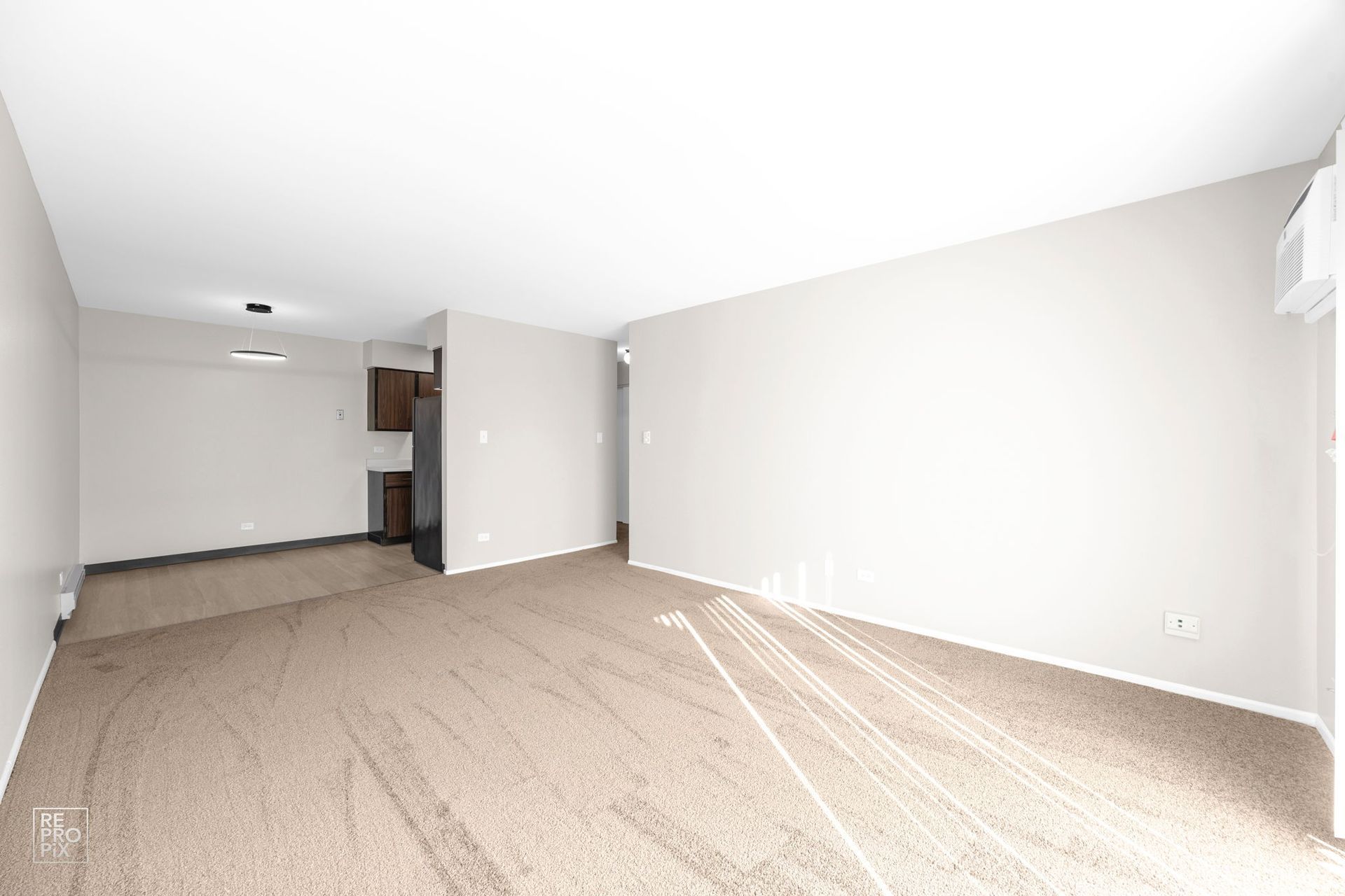 Empty living room with tan carpet and beige walls, with a glimpse of the kitchen.