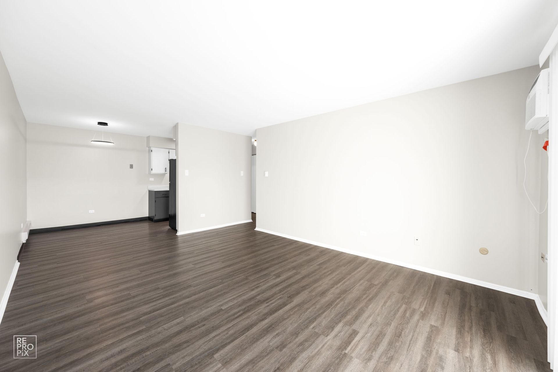 Empty room with dark gray wood-look flooring, white walls, and a small kitchen area.