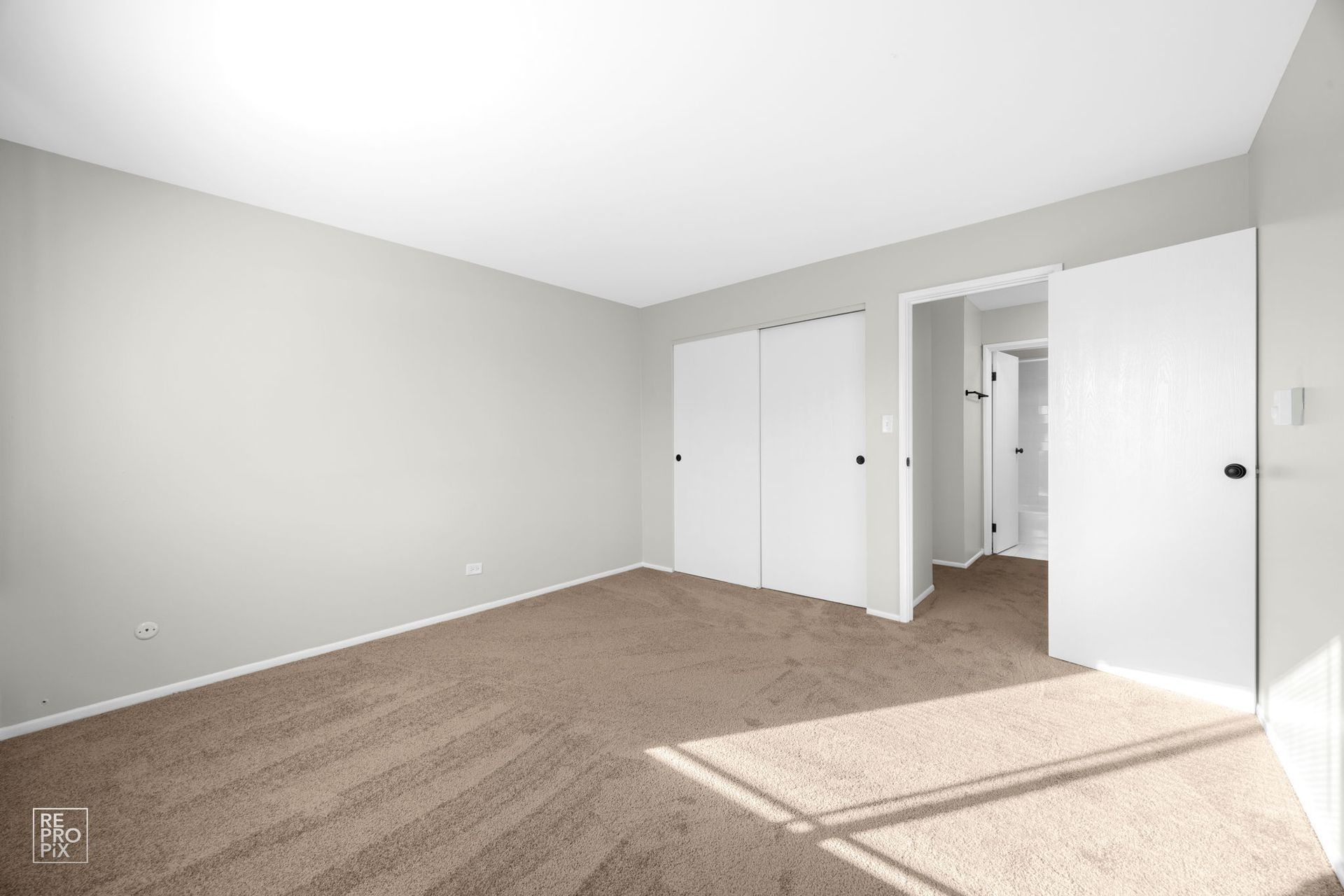 Empty bedroom with beige carpet, light gray walls, white closet doors, and an open doorway.