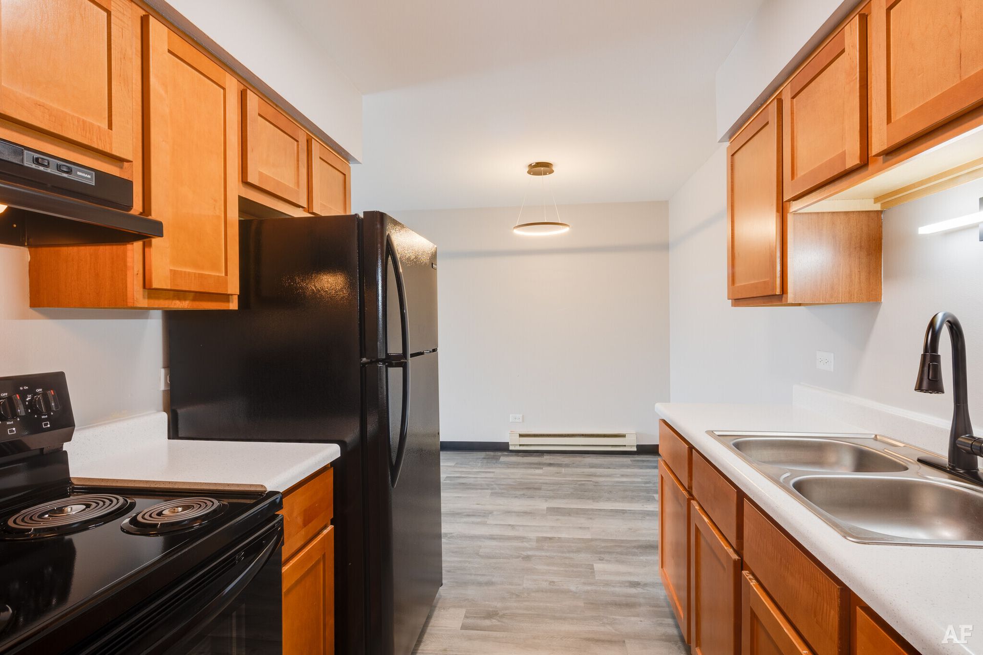 Kitchen with brown cabinets, black appliances, white countertops, and gray flooring; doorway into a light-filled room.