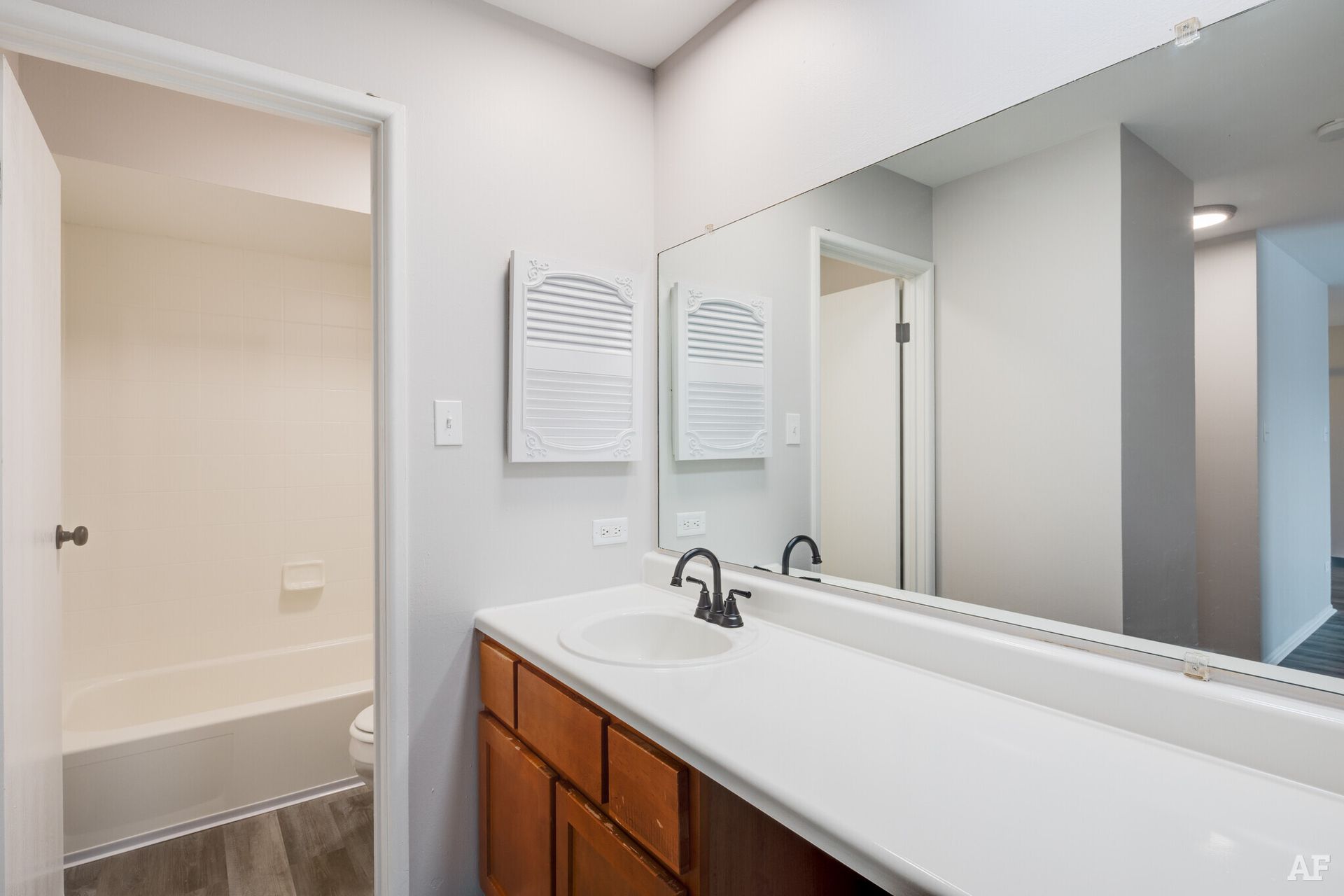Bathroom with a long mirror, wooden cabinets, and a doorway to a bathtub area.