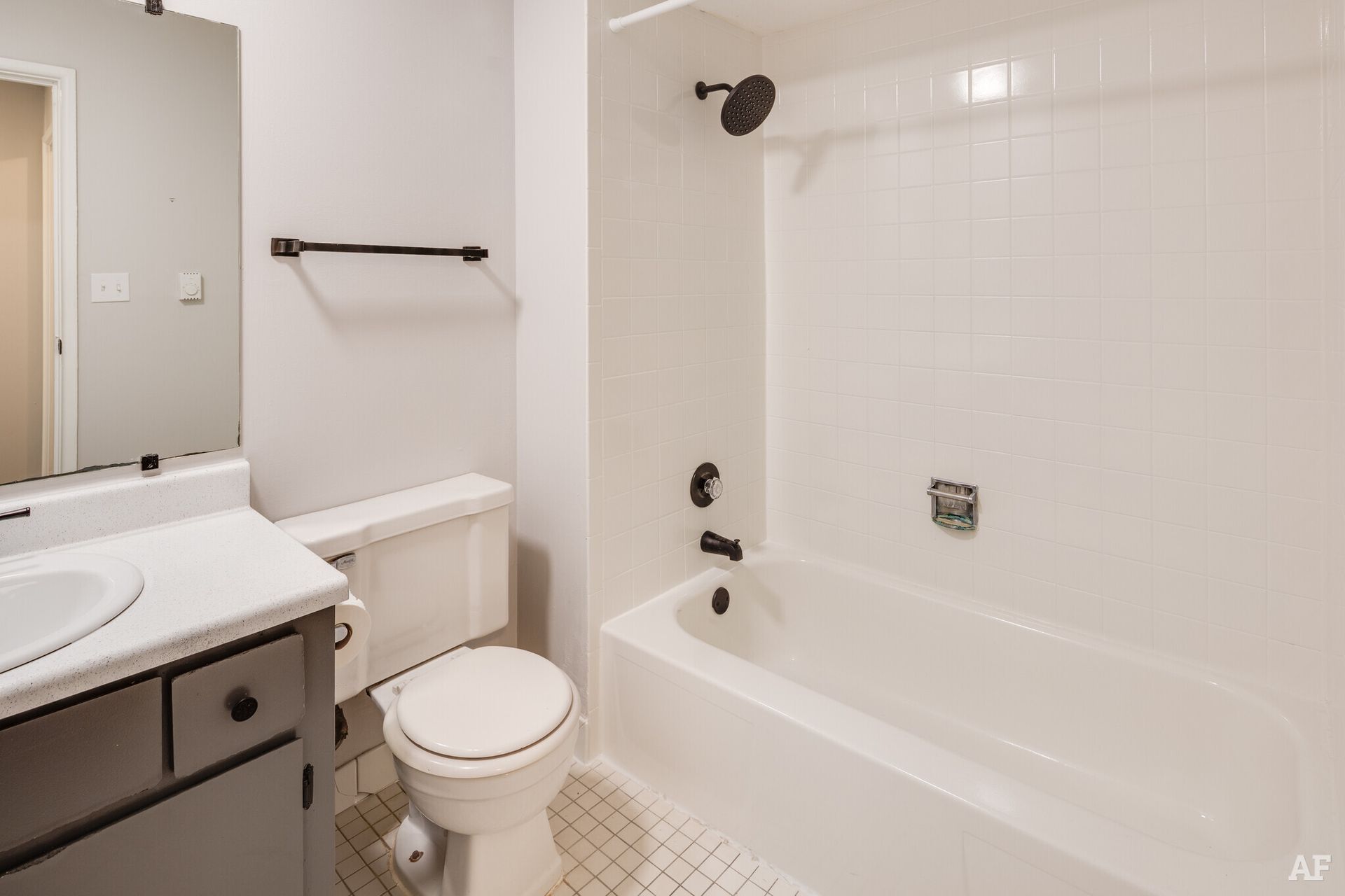Bathroom with white fixtures, light gray walls, dark hardware, and a small, patterned tile floor.