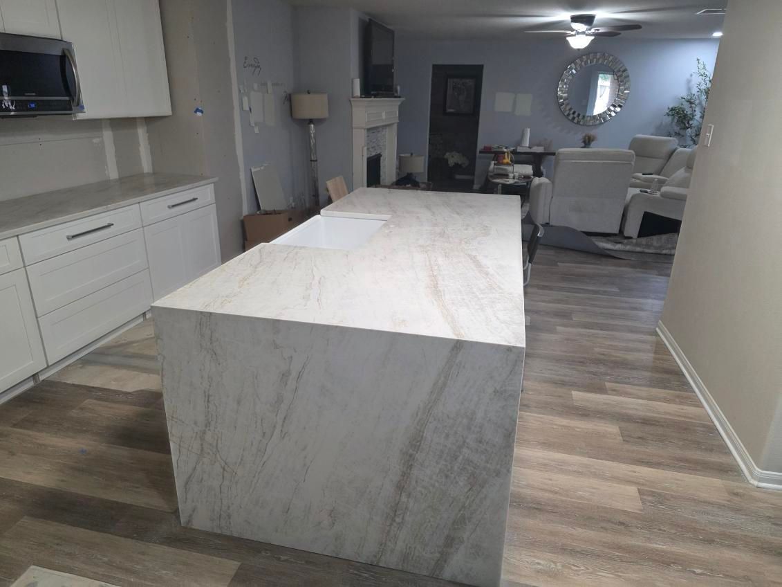 Kitchen with large marble-look island, white cabinets, and living area in the background with wood-look flooring.