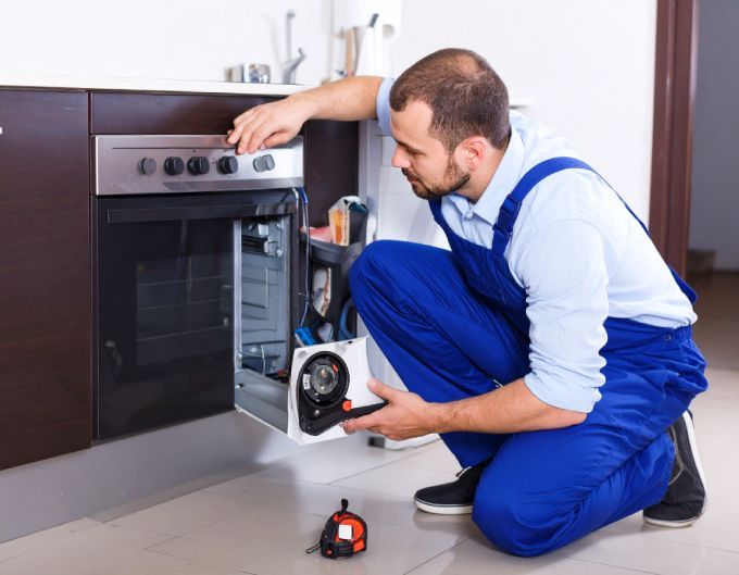 Man in blue overalls repairs an oven in a kitchen. He is crouched down, working on the appliance.