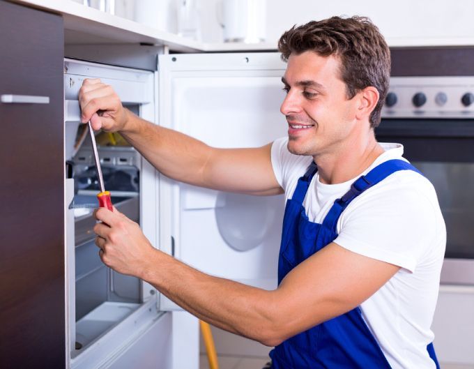 Man in blue overalls smiling, repairing a refrigerator with a screwdriver. Kitchen setting.
