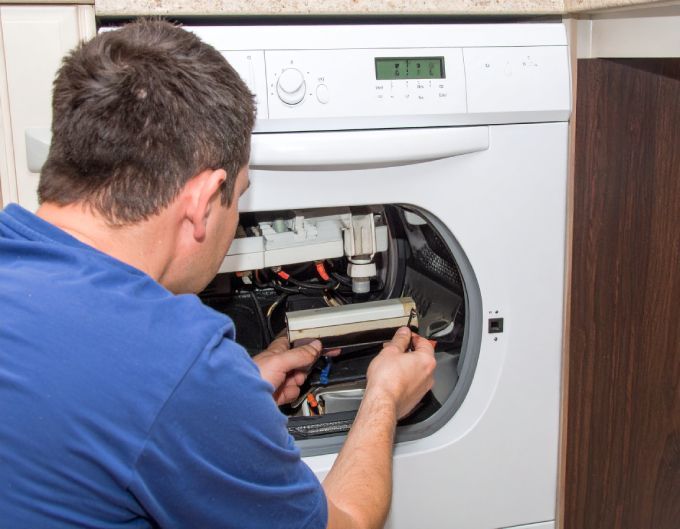 Man in blue shirt repairing a washing machine, examining internal components in a kitchen setting.