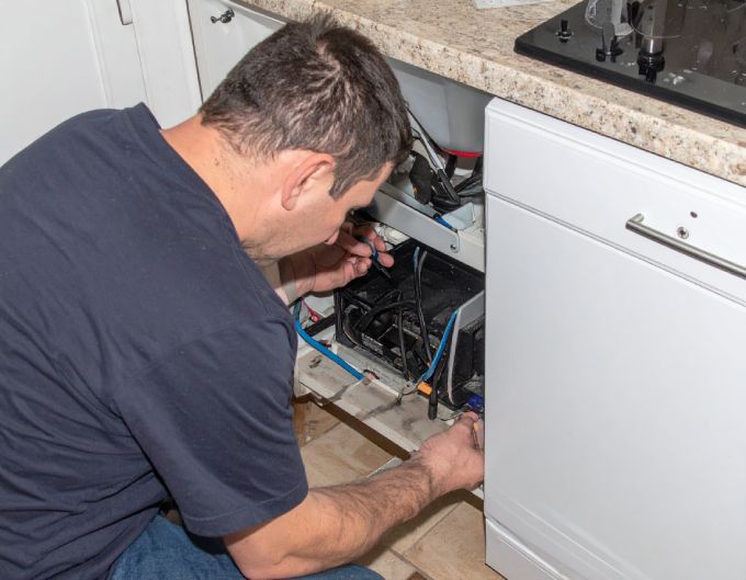 Man repairing a dishwasher in a kitchen, crouched down, focused on wiring.