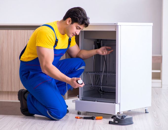 Repairman in yellow shirt and blue overalls fixing a white dishwasher in a kitchen.