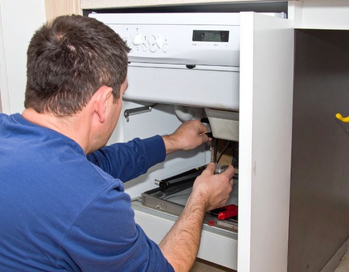 Man in blue shirt fixing a white oven. Working on components inside oven cabinet.