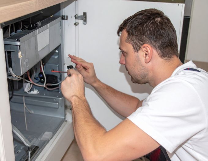 Man in white shirt repairing appliance with a screwdriver; indoor, kitchen setting.
