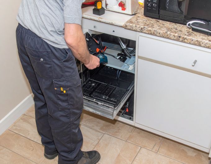A repairman in a gray shirt and blue pants, fixing a dishwasher in a kitchen.