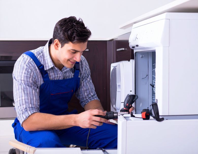 A man in blue overalls repairs a white appliance with a drill in a kitchen.