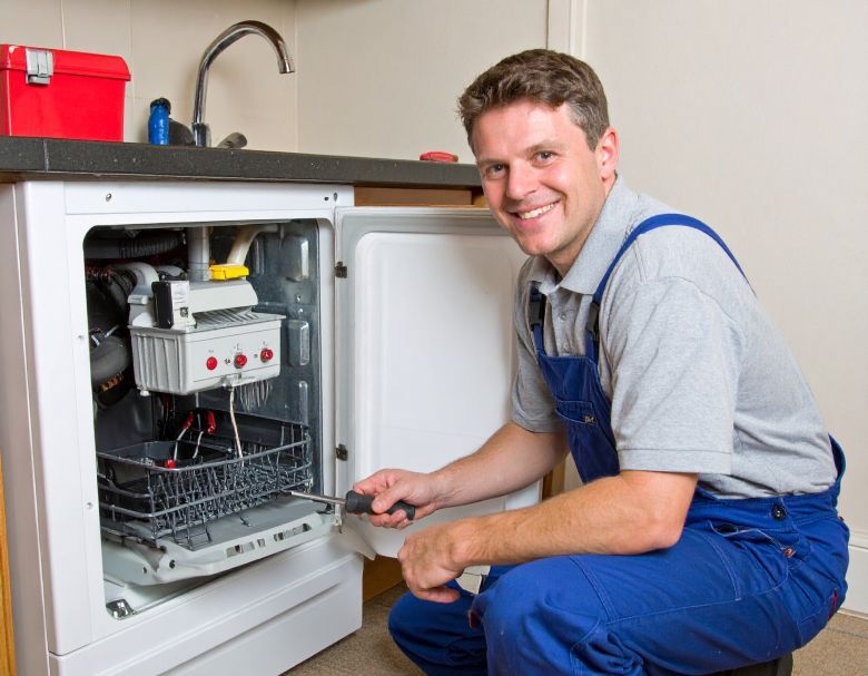 Man in blue overalls repairing a dishwasher, smiling. Kitchen setting.