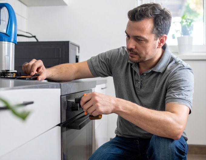Man repairs oven, kneeling in kitchen, gray shirt, focused expression.