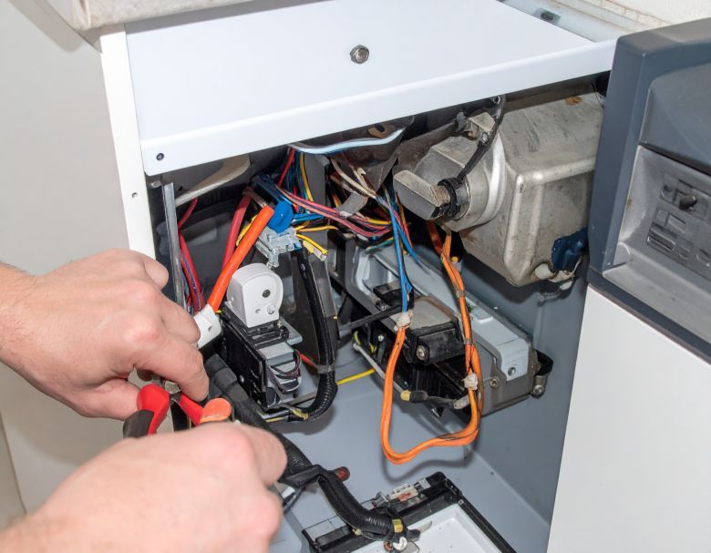 Hands using pliers on the inside of a furnace, exposing wires and components; white and gray tones.