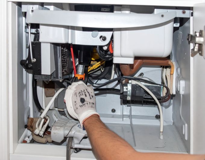 Person in a white-gloved hand working on the interior of a white appliance cabinet with wires, plumbing.