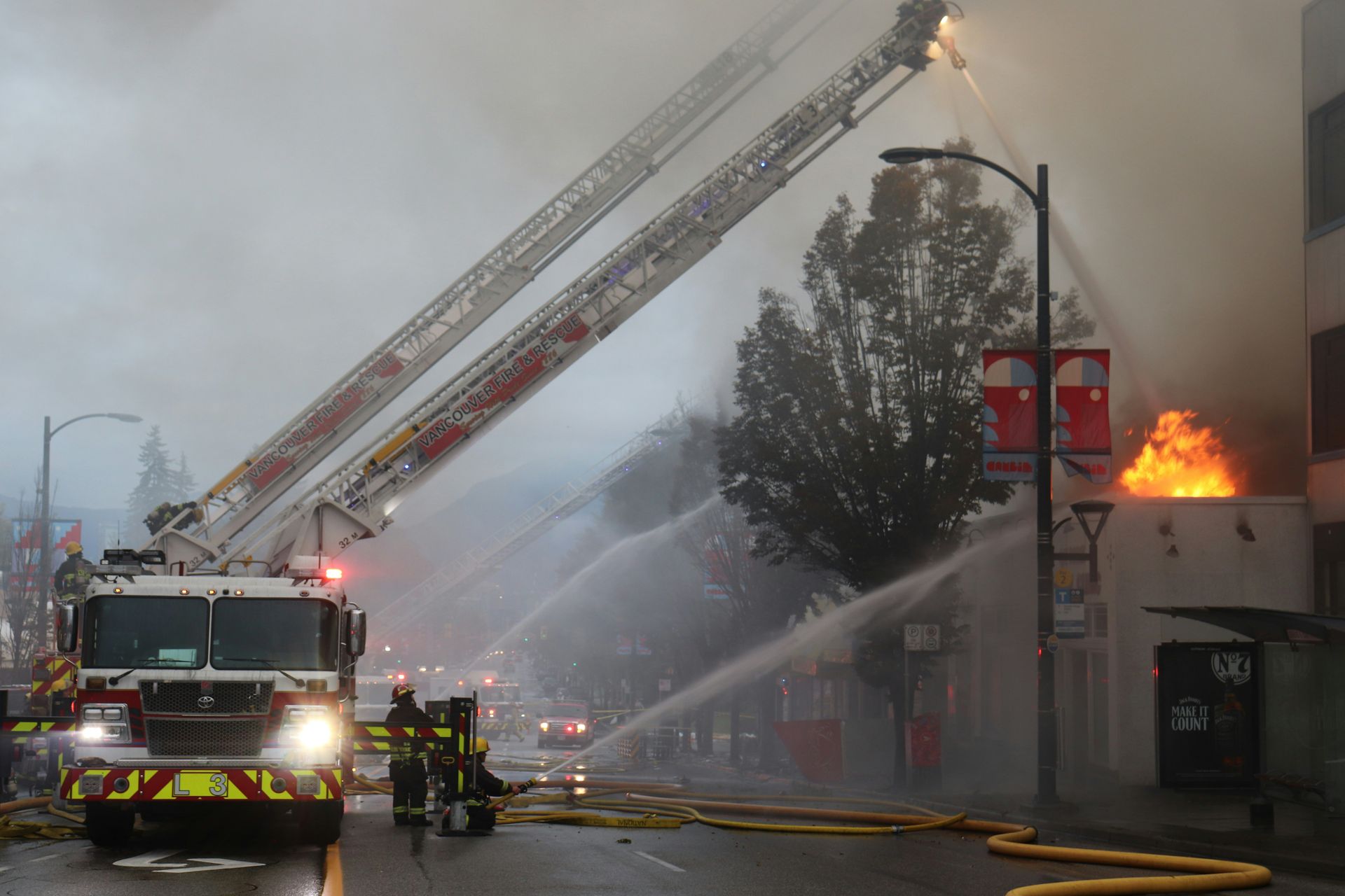 Firefighters battling a building fire. Two ladder trucks spray water; smoke fills the air.