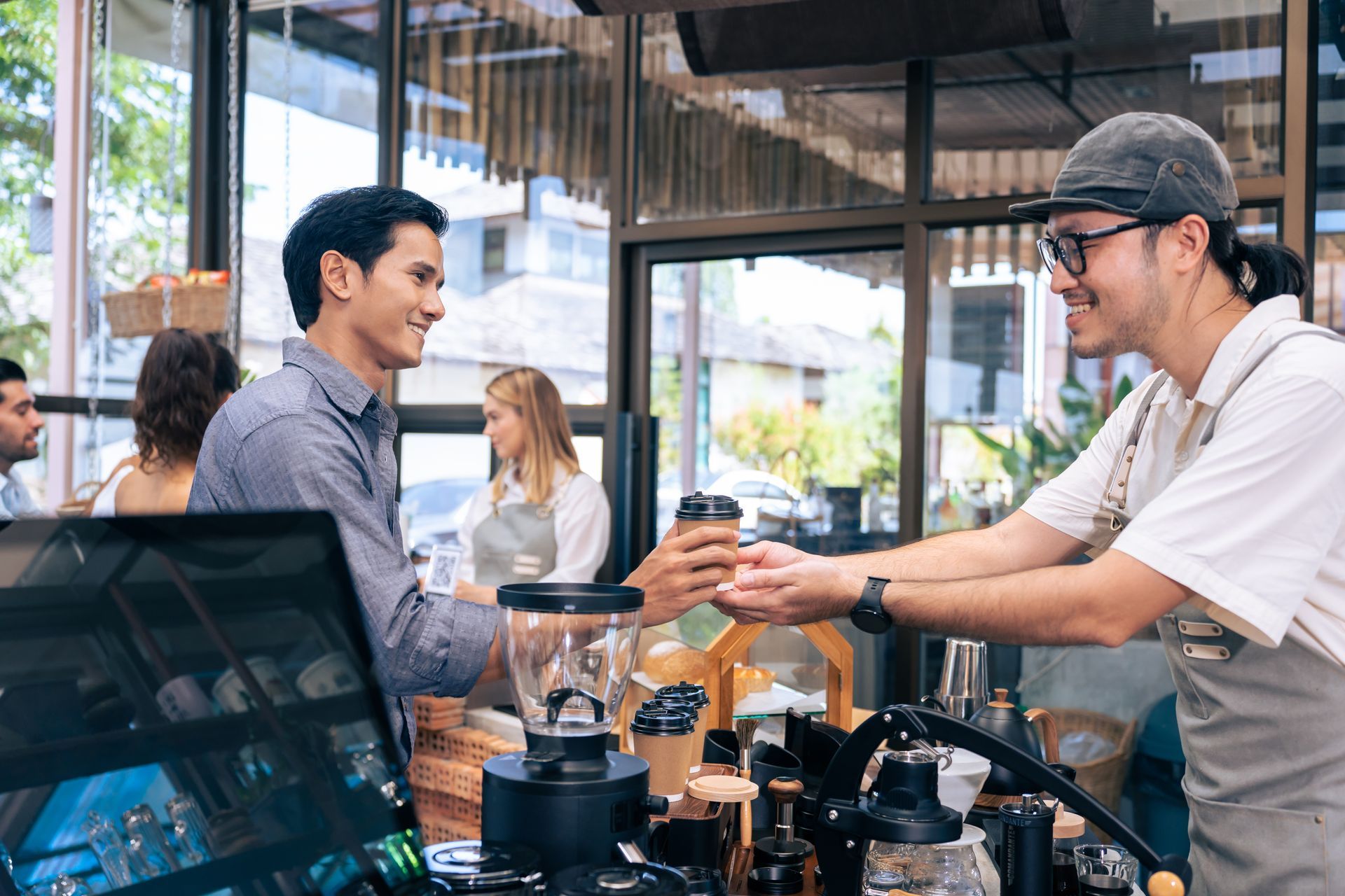 Man receiving coffee from barista at a cafe counter. Other customers and barista in background.