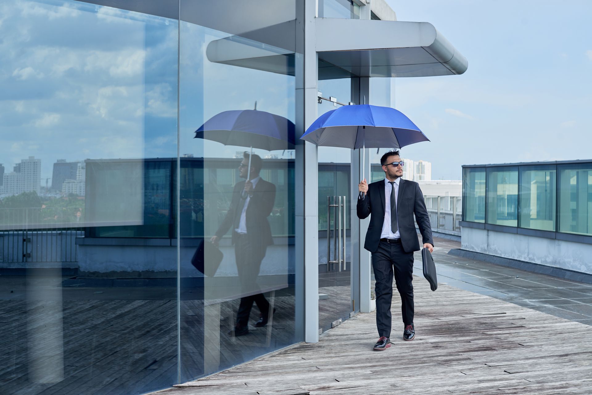 Man in suit holding umbrella and briefcase walking on a rooftop next to glass wall.