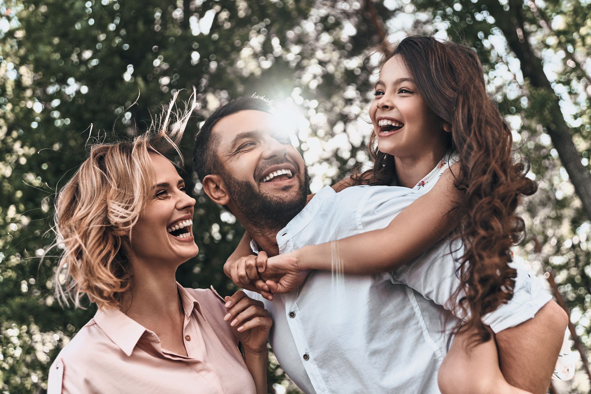 Family laughing, outdoors. Man carries child on his back; woman smiles. Sunlight through trees.