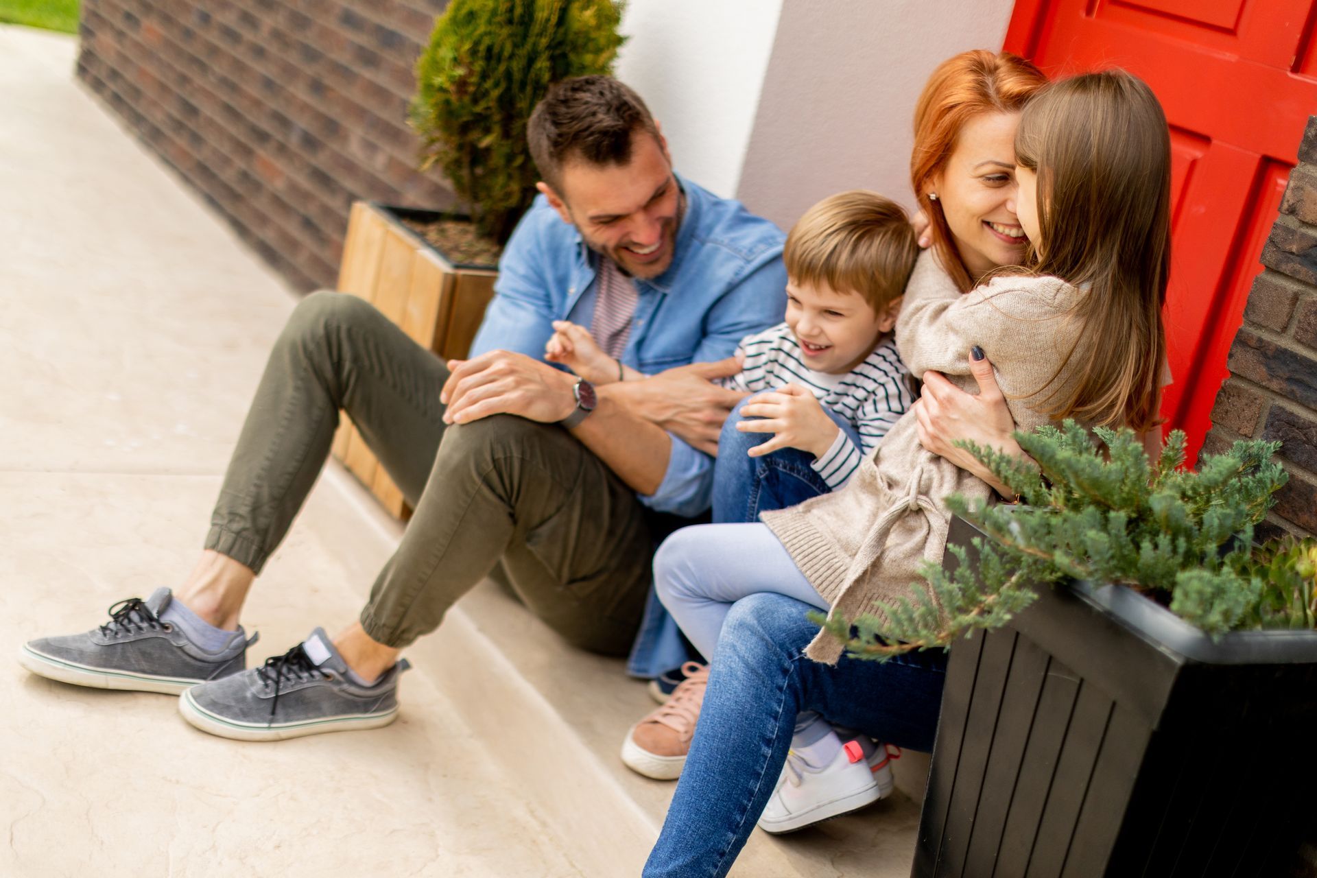 Family of four sitting on steps by a red door; smiling.