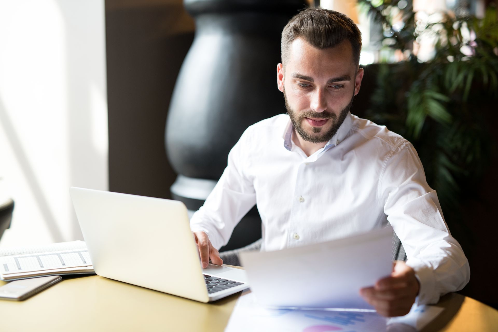 Man in white shirt looking at papers and laptop at a desk.