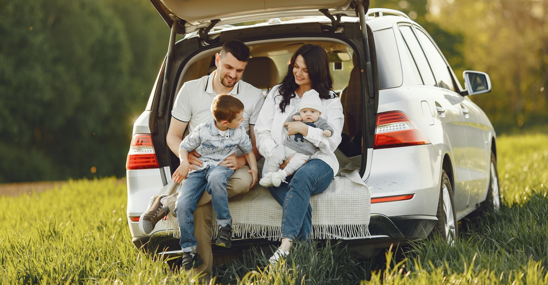 Family sitting in the open trunk of a car in a grassy field.