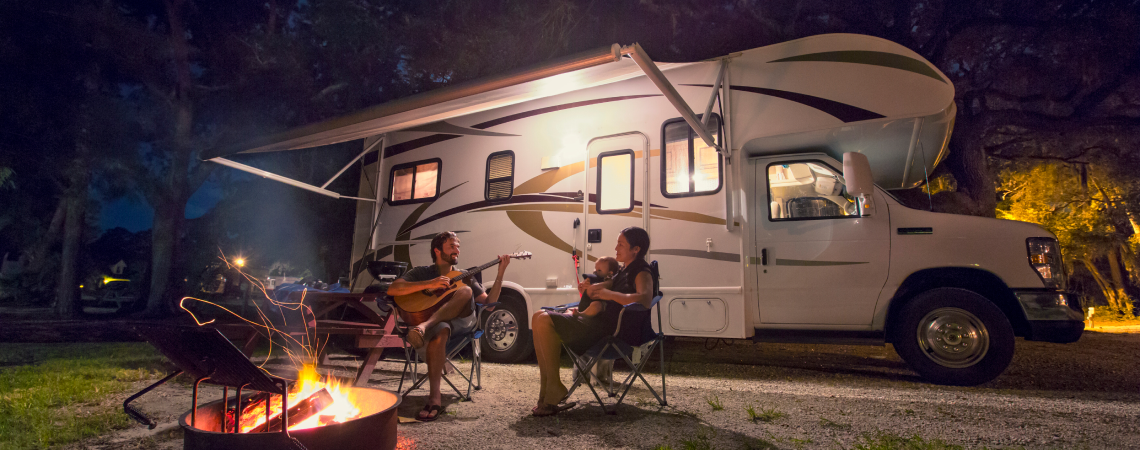 Two people play guitars by a campfire next to an RV at night.