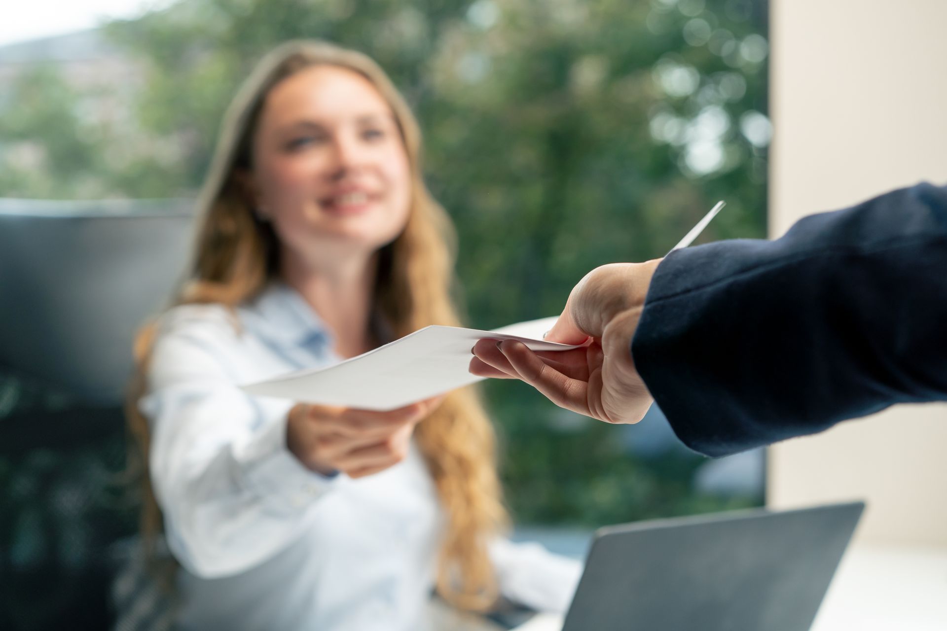 Person in suit hands papers to smiling woman at a desk in an office.
