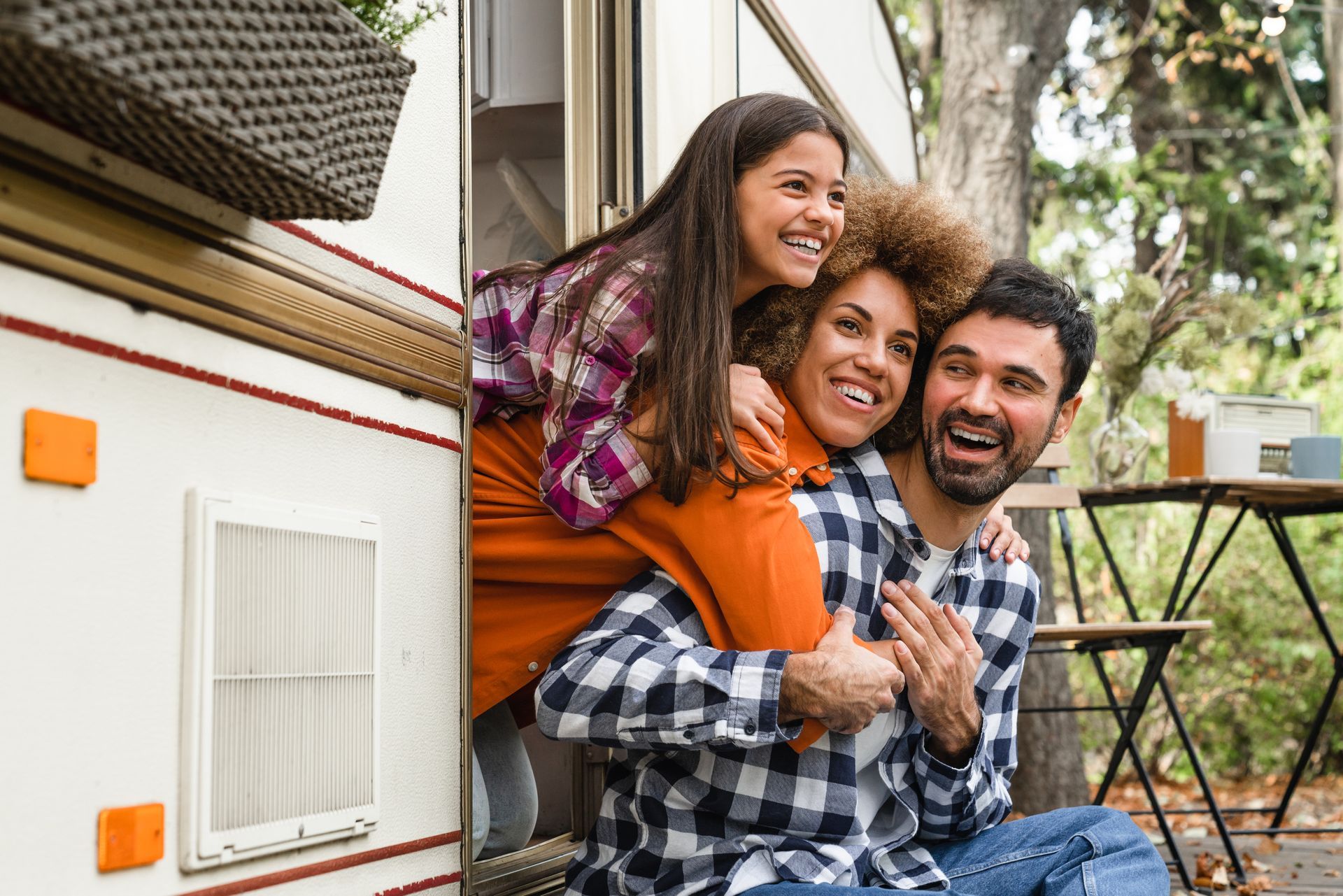 Family of three smiling, posing by a camper in a wooded area.