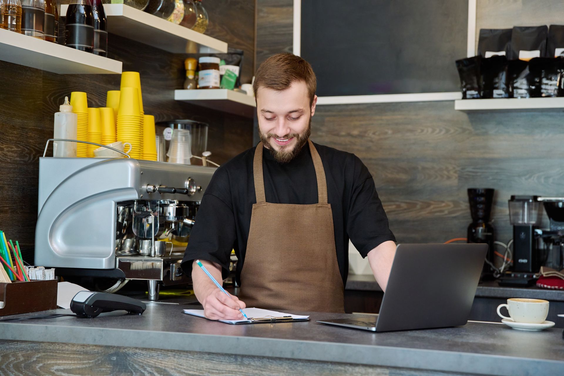 Barista in apron, smiling, working at counter with laptop, taking notes, espresso machine visible.