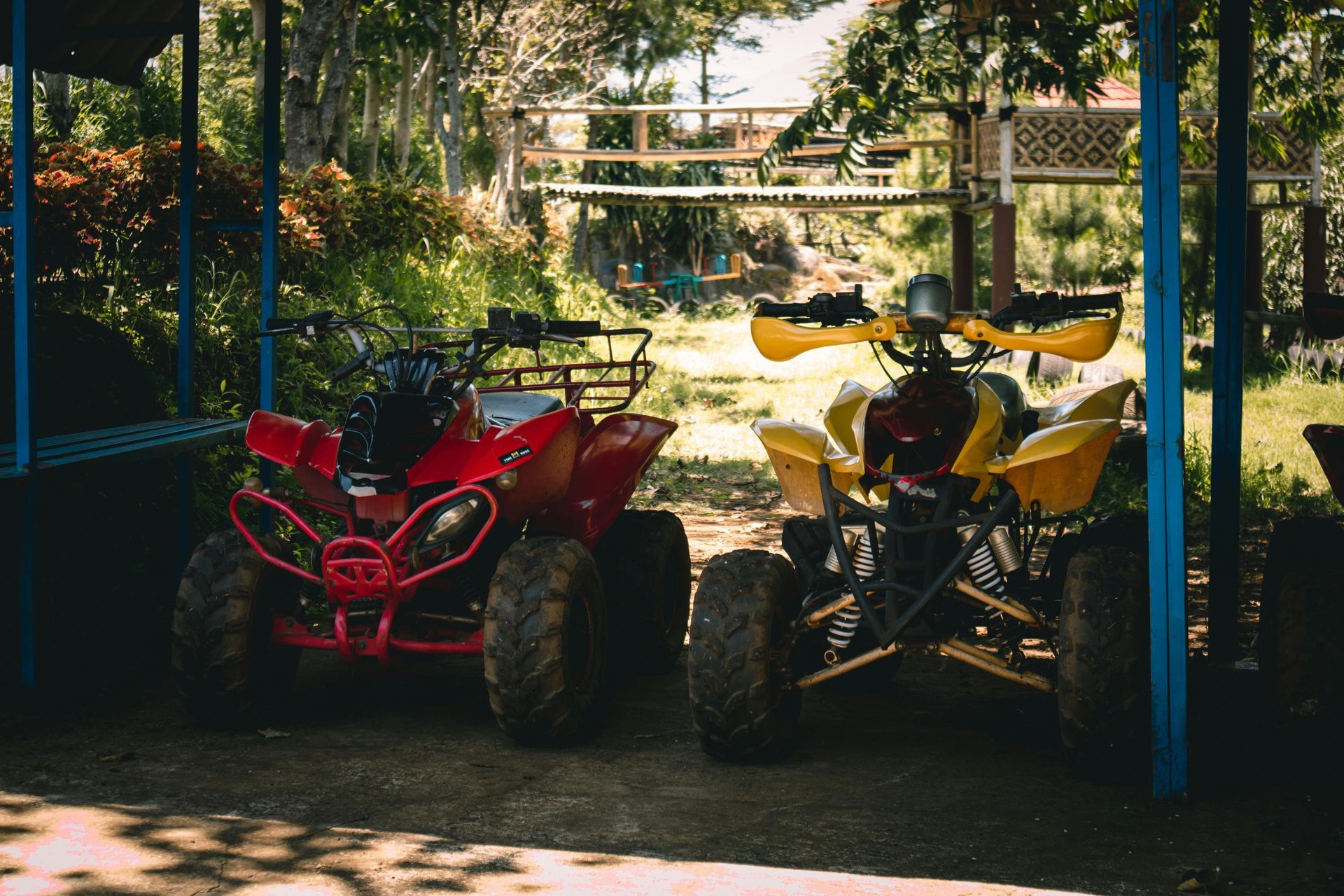Red and yellow ATVs parked under a blue-framed shelter, with a wooden bridge in the background.